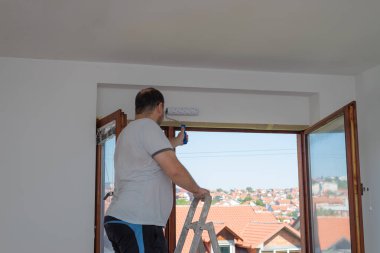 Merdivendeki adam pencerenin üstündeki duvarı beyaz renkte silindirle boyuyor. Pencere açık ve kırmızı çatılar görülebiliyor..