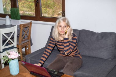 Cep telefonuyla konuşan bir kadın, oturma odasında otururken bilgisayarla çalışan bir kadın.