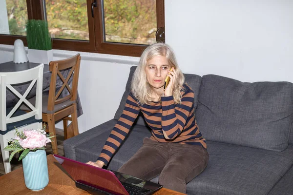 Cep telefonuyla konuşan bir kadın, oturma odasında otururken bilgisayarla çalışan bir kadın.