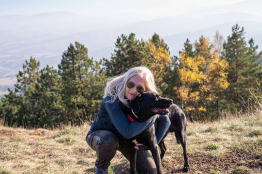 Sarışın bir kadın güneşli bir günde dağda siyah köpeğini kucaklıyor.