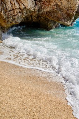  Gök mavisi dalga, kayaların arkasındaki kumu kaplıyor.                                   