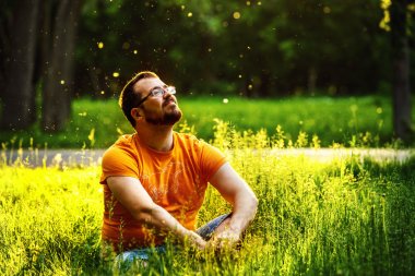 A happy thoughtful dreamer man is sitting on green grass in a park at sunny summer day and looking into future. Concept of relaxation, wellbeing, lifestyle.