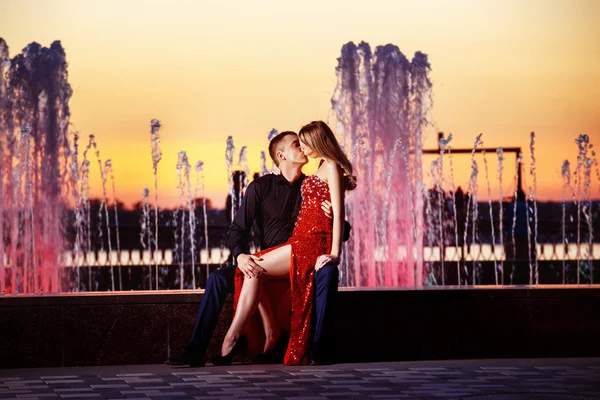 Beautiful young woman in red dress and elegant man kissing on colorful fountain at golden stunning sunset background.