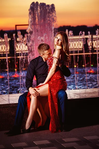 Beautiful young woman in red dress and macho man are kissing at colored water fountain at orange sunset sky background.