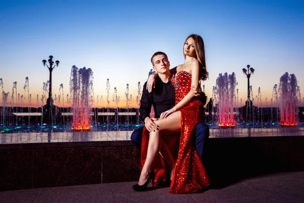 Beautiful young woman in red dress and macho man are sitting at fountain at blue sunset sky background.