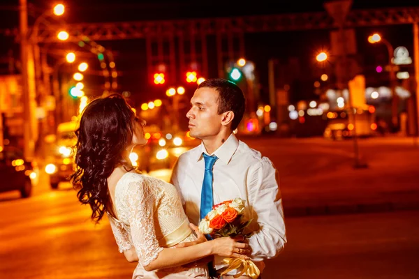 Elegant dresses man is tenderly embracing happy smiling woman with flowers bouquet at bright night street lights background.