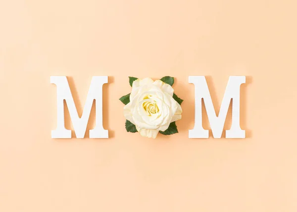 Minimal mom word  made of white letters and soft rose flower on pastel powder background. Trendy mothers day concept.