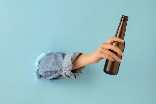 Main féminine avec bouteille de bière brise à travers fond de papier bleu. — Photo
