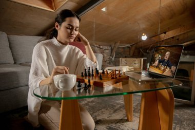 Genç beyaz kız evde oturuyor ve bilgisayarla online satranç oynuyor..