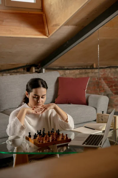 Молодая красивая девушка сидит дома и играет в шахматы онлайн с помощью ноутбука. — стоковое фото