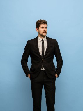Siyah takım elbiseli yakışıklı genç adam mavi arka planda yalnız poz veriyor..