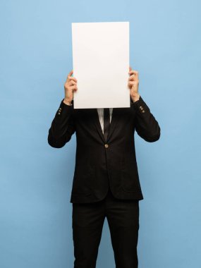 Siyah takım elbiseli, mavi arka planda boş bir kağıt parçası olan genç bir adam..