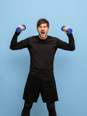Mavi arka planda el ağırlıkları olan siyah spor üniformalı genç bir adam..