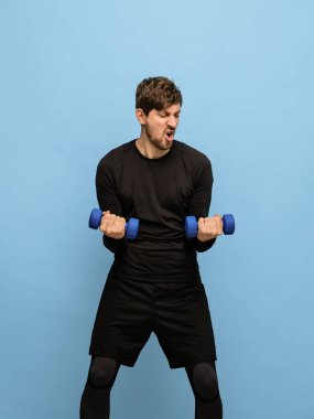 Mavi arka planda el ağırlıkları olan siyah spor üniformalı genç bir adam..