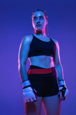 Atletik bayan MMA dövüşçüsü mavi arka planda, neon ışıkta yalnız poz veriyor.