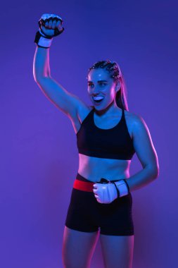 Genç bayan MMA dövüşçüsü mavi-pembe arka planda, neon ışıkta yalnız poz veriyor.