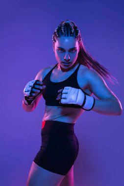 Atletik bayan MMA dövüşçüsü mavi arka planda, neon ışıkta yalnız poz veriyor.