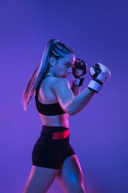 Genç bayan MMA dövüşçüsü mavi-pembe arka planda, neon ışıkta yalnız poz veriyor.