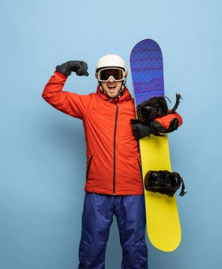 Parlak kayak takımı ve snowboard giymiş genç adam mavi arka planda yalnız poz veriyor..