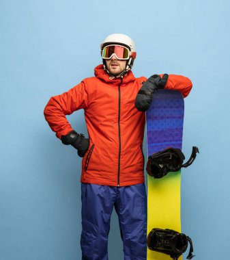 Mavi arka planda parlak kayak takımı ve snowboard giyen genç adam izole edilmiş..