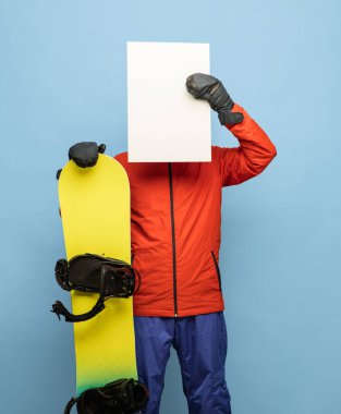 Parlak kayak takımı ve snowboard giymiş genç adam mavi arka planda yalnız poz veriyor..