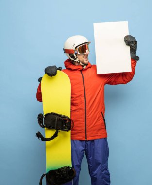 Parlak kayak takımı ve snowboard giymiş genç adam mavi arka planda yalnız poz veriyor..