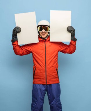 Üzerinde işaret olan parlak kayak takımlı genç bir adam, mavi arka planda poz veren bir kağıt.