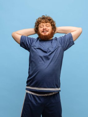 Mavi spor üniformalı genç, kızıl saçlı adam mavi arka planda izole edilmiş..