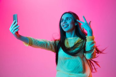 Genç güzel bir kız pembe arka planda neon ışıkta izole edilmiş selfie çekiyor.