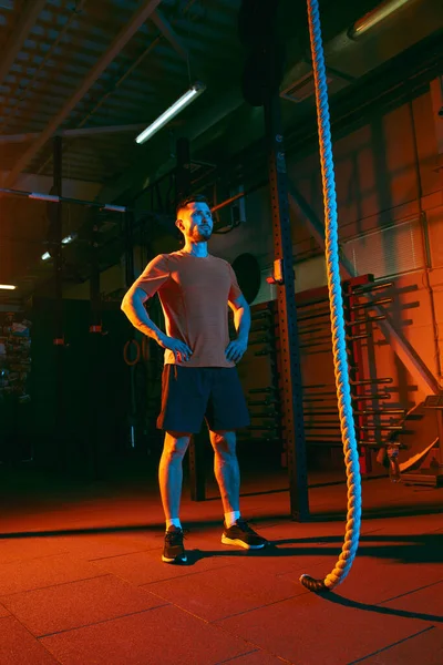 Young athletic man, fitness instructor training in gym in mixed neon light.