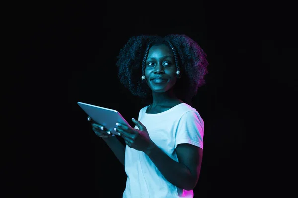 Jong Afro-Amerikaans meisje met behulp van tablet geïsoleerd op donkere achtergrond in neon. — Stockfoto