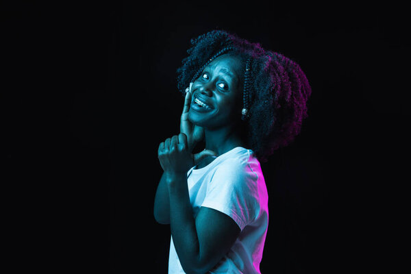 Stylish hairdo. Half-length portrait of young beautiful African-American girl isolated on dark background in multicolored neon light. Concept of human emotion, facial expression. Copy space for ad
