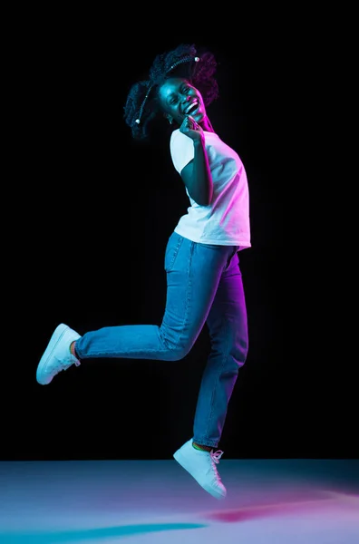 Giovane ragazza afroamericana che salta isolata su sfondo scuro al neon. — Foto Stock