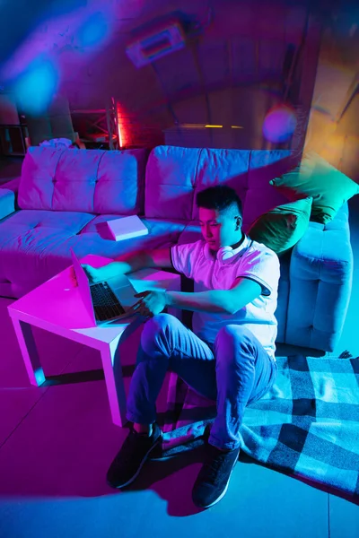 Young Korean man using laptop at home interior in gradient pink blue neon light