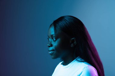 Gülümseyen genç Afrikalı güzel bir kız karanlık arka planda, karışık neon ışıkta izole edilmiş. Yan görünüm