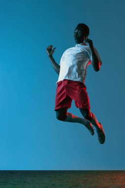 Genç Afrikalı futbolcu mavi arka planda neon ışıkta tek başına zıplıyor.