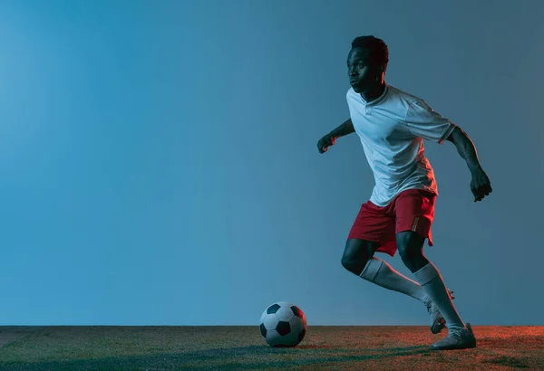 Junger afrikanischer Fußballspieler springt isoliert auf blauem Hintergrund im Neonlicht — Stockfoto