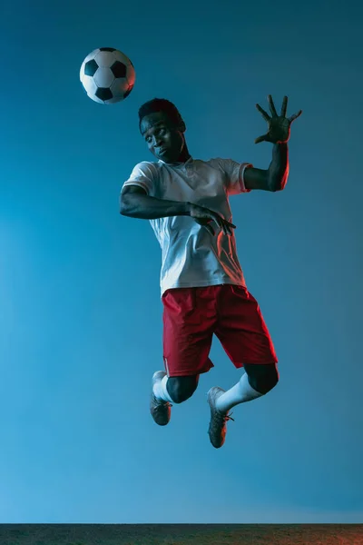 Νεαρός αφρικανός ποδοσφαιριστής που πηδά απομονωμένος σε μπλε φόντο υπό νέον φως — Φωτογραφία Αρχείου
