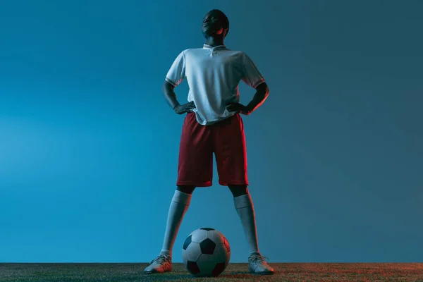 Νεαρός αφρικανός ποδοσφαιριστής που ποζάρει απομονωμένος σε μπλε φόντο υπό νέον φως — Φωτογραφία Αρχείου