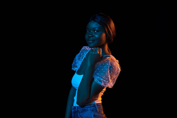 Calm, smiling. Portrait of beautiful African girl isolated on dark background in neon mixed light. Female model in casual clothes. Concept of human emotions, facial expression, youth. Copyspace for ad
