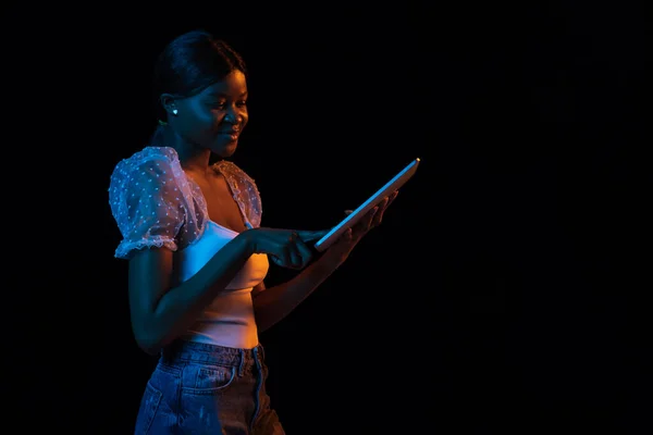 Молодая африканская красивая девушка с планшетом изолированы на темном фоне в смешанном неоновом свете. — стоковое фото