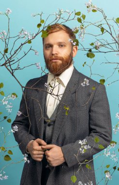 Genç kızıl saçlı ve sakallı bir adamın portresi. Ünlü bir sanatçıymış gibi davranıyor. Mavi çiçekli arka planda izole edilmiş.