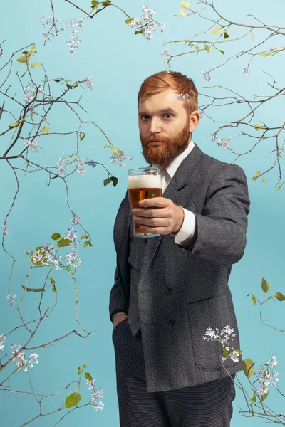 Genç kızıl saçlı ve sakallı bir adamın portresi mavi çiçekli arka planda izole edilmiş bir bardak birayla