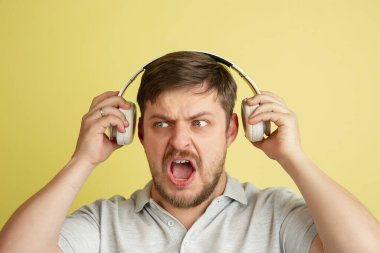 Kulaklıklı genç bir adam açık sarı arka planda izole edilmiş. Yüz ifadeleri, insan duyguları kavramı.
