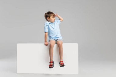 Gri stüdyo arka planında izole edilmiş, günlük giysiler içinde, kutunun üzerinde oturan beyaz bir çocuk..