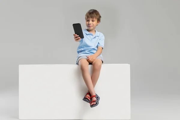 One Caucasian little boy in casual clothes sitting on box isolated over gray studio background. — Stock Photo, Image