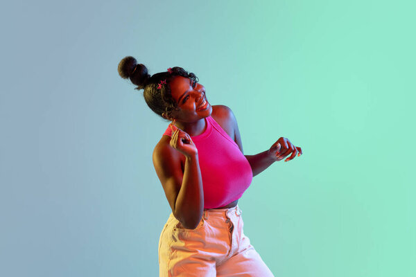 Joyful young African girl, female fashion model dancing at camera isolated on blue green background in neon light. Concept of human emotion, facial expression. Copy space for ad.