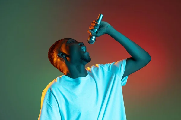 Neon gradyan arka planda mikrofonla şarkı söyleyen Afrikalı genç bir adam. Duygu, eylem, yüz ifadesi kavramı. — Stok fotoğraf