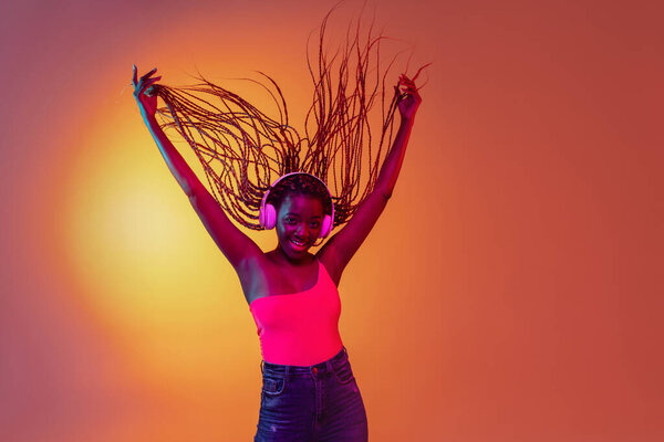 Happiness. Young pretty flighty African girl with afro braids in white headphones isolated on yellow background in neon light. Concept of human emotion, facial expression.