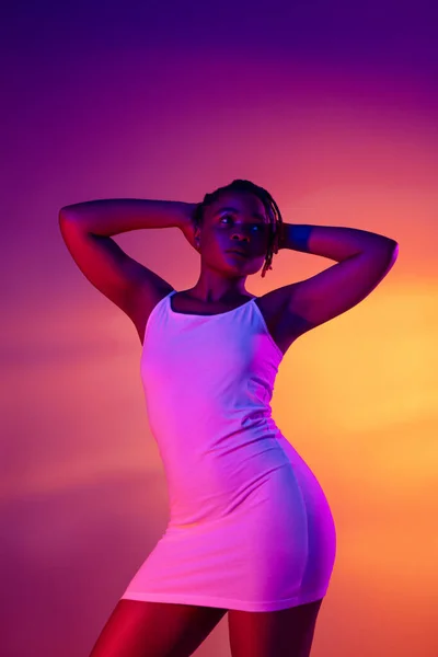Vacker afrikansk flicka i vit kort sommar klänning poserar isolerad över ljusa färgglada neon bakgrund. — Stockfoto